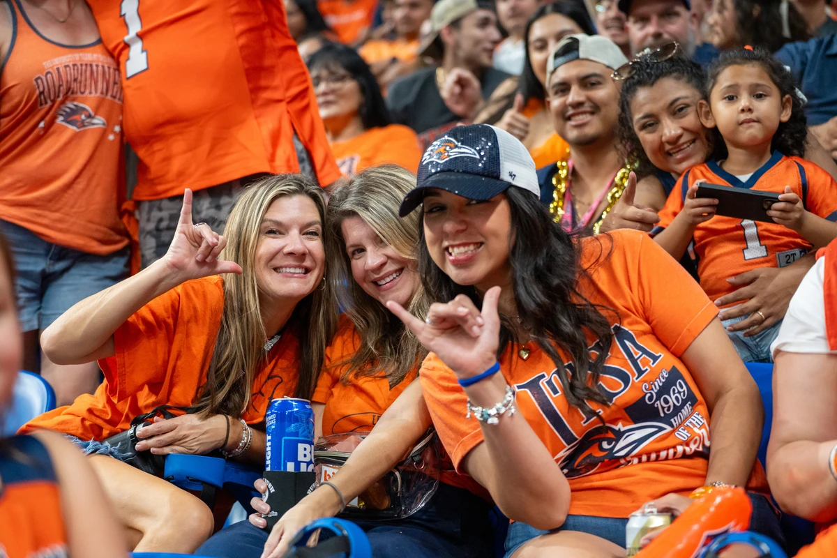 three women in a crowd of peopole wearing orange UTSA T-shirts in a stadium, smiling and holding up the "birds up" hand sign.